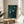 May include: Framed artwork featuring three white birds in flight against a dark teal-green textured background, displayed on a round wooden side table next to a beige upholstered chair. Natural sunlight streams across the white wall, casting shadows from a plant's leaves. The piece has a minimalist Asian-inspired aesthetic with vertical brushstroke textures and small red signature stamps in the lower corner.