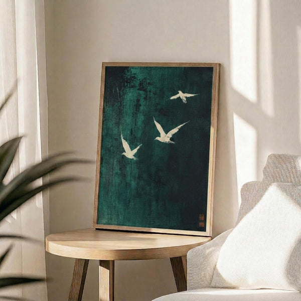 May include: Framed artwork featuring three white birds in flight against a dark teal-green textured background, displayed on a round wooden side table next to a beige upholstered chair. Natural sunlight streams across the white wall, casting shadows from a plant's leaves. The piece has a minimalist Asian-inspired aesthetic with vertical brushstroke textures and small red signature stamps in the lower corner.