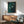 May include: Framed artwork with green background and white birds in flight, displayed above a wooden credenza in modern living room setting. Mid-century style furniture includes green upholstered armchair with wooden frame and small table lamp. Natural sunlight creates shadows on white wall.