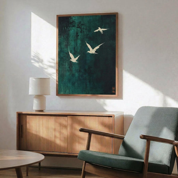May include: Framed artwork with green background and white birds in flight, displayed above a wooden credenza in modern living room setting. Mid-century style furniture includes green upholstered armchair with wooden frame and small table lamp. Natural sunlight creates shadows on white wall.