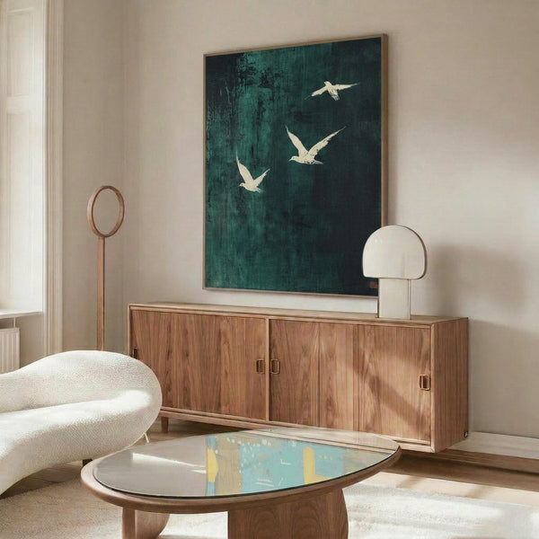 May include: Modern living room with wooden credenza displaying large framed artwork featuring three white birds flying against dark teal-green textured background. Scene shows white curved chair, oval glass coffee table, mushroom-style table lamp, and circular floor mirror with wooden frame on light walls.