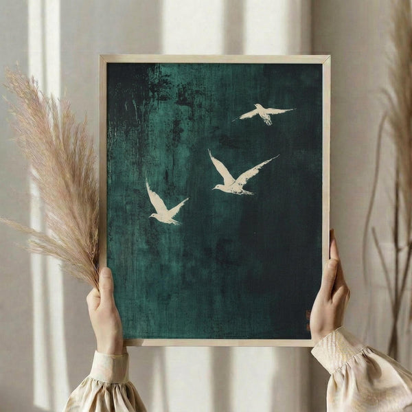 May include: Person holding framed artwork showing three white birds in flight against dark teal-green textured background, displayed in light wooden frame. Dried pampas grass visible on left side. Soft natural lighting from window creates gentle shadows on neutral wall behind.