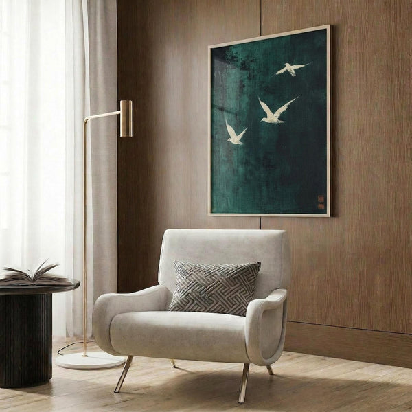 May include: A framed teal and dark green artwork featuring three white birds in flight, displayed on a wood-paneled wall above a beige upholstered armchair with geometric pillow. The modern room setting includes a brass floor lamp and dark round side table with an open book.