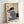 Serene framed poster of a geisha applying makeup at her mirror, executed in the Japanese woodblock style with a refined palette of cream, pale blue, and deep indigo.