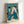 Introspective portrait of a geisha with mirror in a classical Japanese woodblock aesthetic, combining muted cream and black with rich teal blue and ochre highlights.