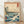Japanese woodblock seascape print featuring Mount Fuji rising majestically above rolling indigo waves, with a pine tree, boat, and birds in muted teal, cream, and burnt orange tones.