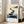 Serene watercolor landscape poster with stepping stones crossing soft teal waters, featuring a sparse tree silhouette and charcoal rock formations in soothing neutral tones.