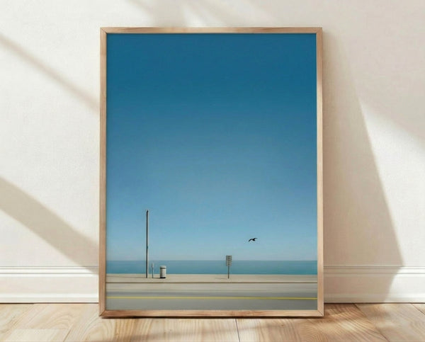 Minimalist coastal landscape framed poster featuring a solitary bird near a trash can and utility pole on a quiet beach road, rendered in soft teal, blue, beige, and grey tones.