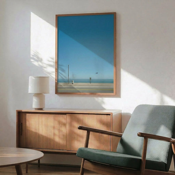 Minimalist beach road poster with contemplative mood, showing a bird and coastal structures under calm skies in gentle blue, grey, and cream tones.
