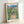 Serene garden composition in soft impressionist style showing sunflowers along a pathway beneath delicate blue sky and green canopy, with figures enjoying a peaceful romantic moment among the blooms.