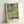 Romantic impressionist poster depicting a wooden Japanese bridge arching above a lily pond, with pale pinks, creams, and gentle greens evoking a dreamy garden landscape.