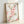 Framed watercolor artwork displaying graceful peach blossoms with layered petals, green foliage, and natural brown branches in soft, romantic palette.
