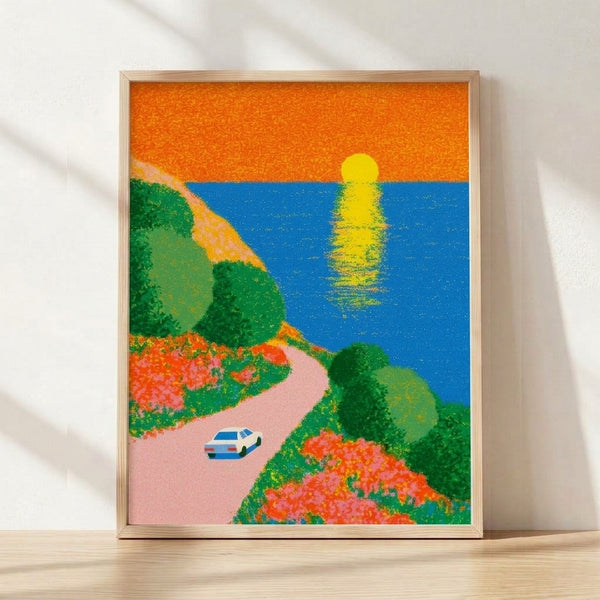 Retro-style framed art print featuring a classic car driving along a coastal cliff road at sunset, with warm orange and pink skies over a blue ocean and green landscape.