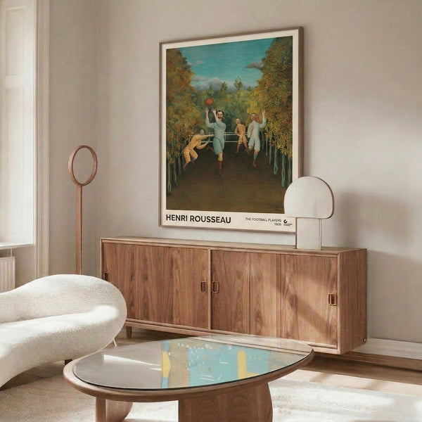 Art print framed on wall showing Rousseau's naive surreal scene of football players amid lush vegetation, rich with cream, forest green, rust, and ochre in charming 1908 composition.
