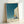 Surreal wall art depicting a solitary stairway rising through a barren landscape with a crescent moon and distant tree, executed in muted teal, grey, and cream with minimalist style.