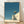 Minimalist surreal artwork depicting a solitary figure ascending stairs toward a crescent moon in a desert landscape, rendered in teal blue, cream, and soft gold tones.