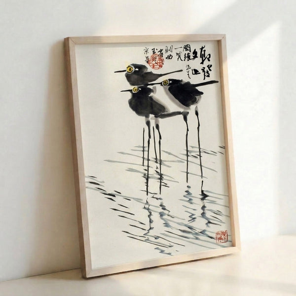 Framed ink wash painting in the style of Japanese folk art showing birds with extended legs in wetland grasses, composed in muted black, gray, and cream palette.