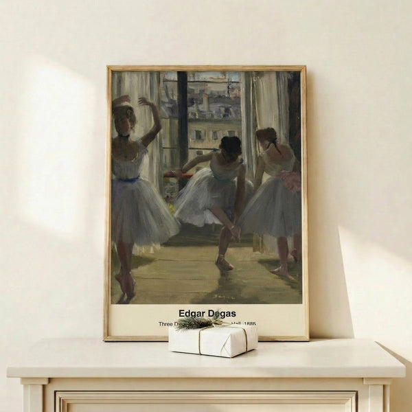 Framed art print capturing three female dancers in an intimate studio setting, executed in pale cream and ochre with subtle blue accents in a classical Impressionist style.