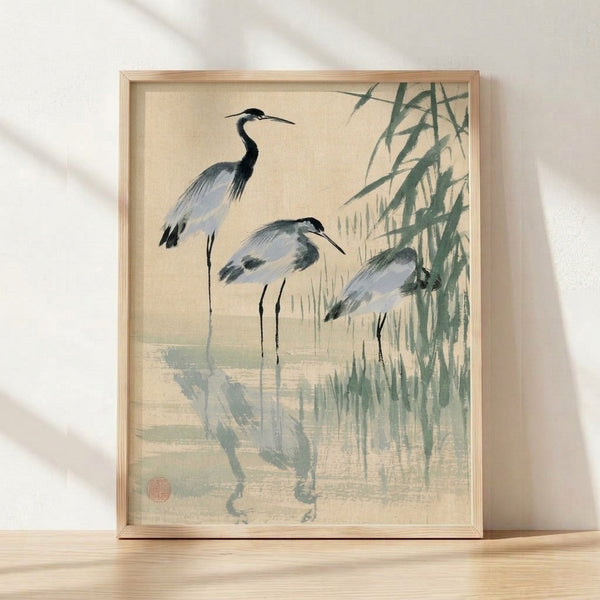 Three herons among tall reeds in a serene Japanese ink wash style, rendered in cream, sage green, and pale blue tones with delicate black brushwork on a framed art print.