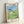 Serene anime poster art showing Totoro amid blooming grassland with an umbrella, rendered in dreamy watercolor-inspired tones of soft green and light blue.