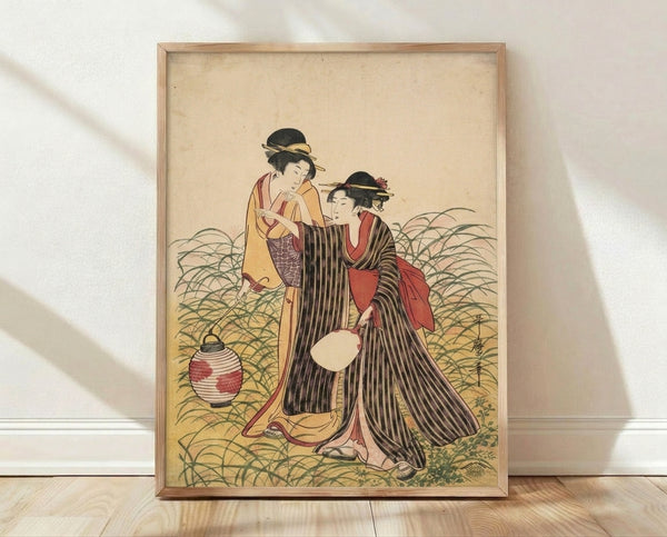 Framed Japanese woodblock print depicting two graceful geisha in ornate kimonos beside a glowing lantern amidst green grass, rendered in cream, black, red, and gold tones with serene elegance.