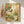 Peaceful watercolor garden scene with a robin surrounded by rose blossoms, potted plants, and a rake, rendered in muted coral pink, sage green, and pale yellow with cottage charm.
