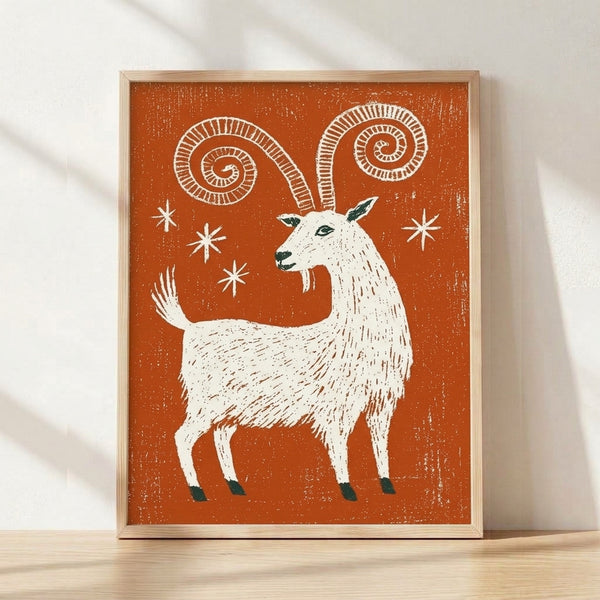 Framed folk art linocut print of a goat with spiral horns and stars, rendered in rust orange, cream, and dark teal on a wall.