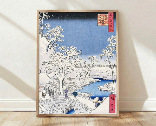 Serene Japanese woodblock print featuring a snow-covered bridge spanning a river with snow-laden trees and distant mountains in deep blue, white, and black tones.