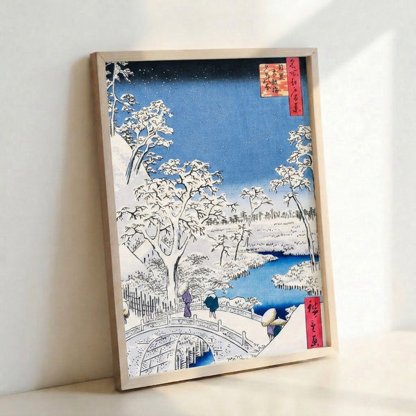 Framed Japanese woodblock art depicting a quiet snow scene with a wooden bridge, forest of bare trees dusted in white, and layered blue mountains beyond.