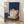 Minimalist Japandi painting of a woman seated by a window holding a coffee cup, surrounded by potted plants and flowers in soft navy, cream, and warm beige tones.