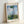 Soft, romantic impressionist scene of a figure in flowing white garment with parasol walking through a blooming field under hazy golden light, framed wall art.